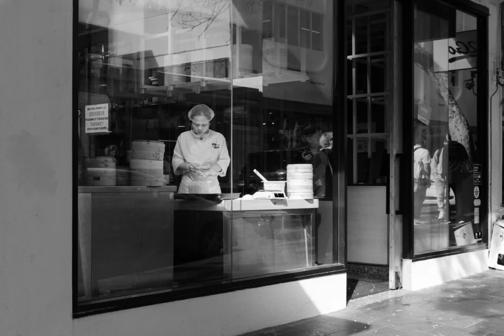 Monochrome image of a chef crafting dumplings by hand through a window. She is framed by a streak of light, while shadows surround the rest of the frame.