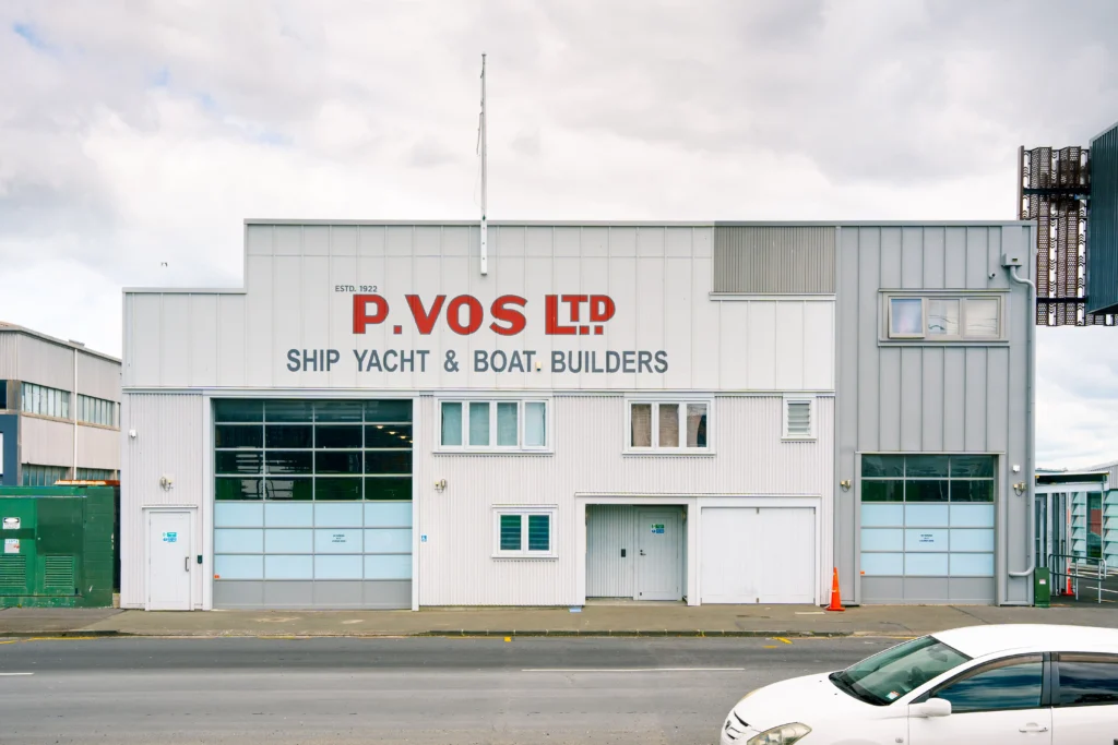 A front on view of the exterior of P. Vos LTD, an old shipyard in Auckland, New Zealand. It's an industrial building, with an empty flag pole, a clear glass garage door and bright red text holding its name.