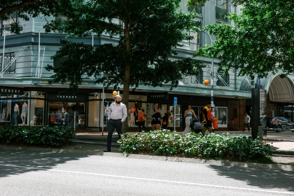 A man illuminated in a beam of sunlight crosses a bustling street in Wellington, NZ. He is surrounded by green trees and shops.