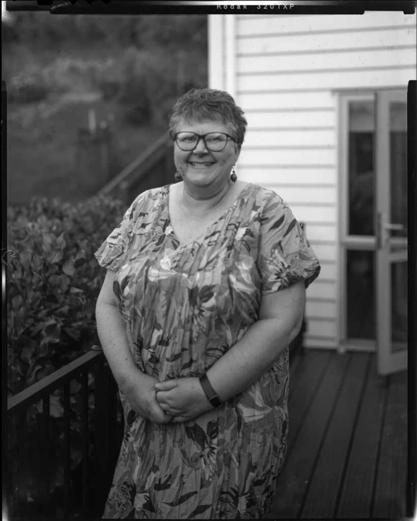 A large format black and white sheet film photograph of a woman standing on the deck of a house. She has short, dark hair and smiles, wearing a flowered dress.
