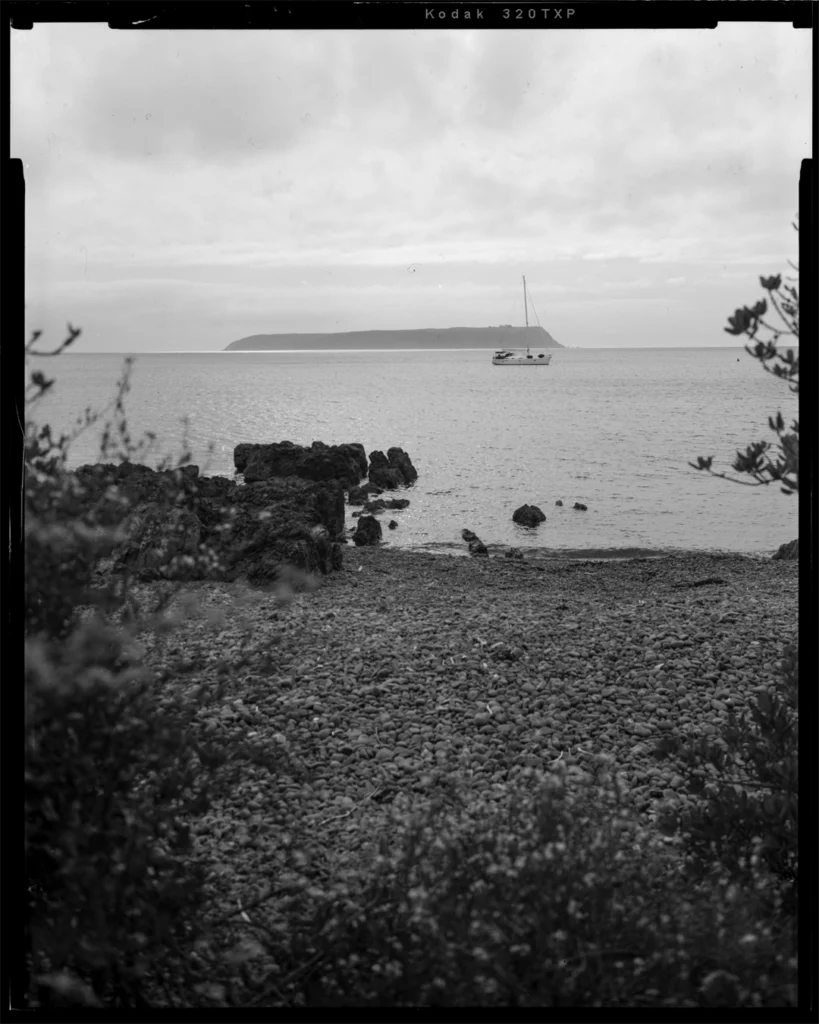 A large format black and white sheet film photograph of an island taken from a rocky beach shore between some foliage. Dark bedrock extends out from the beach, while a small yacht sits idle on the water before the distant island.