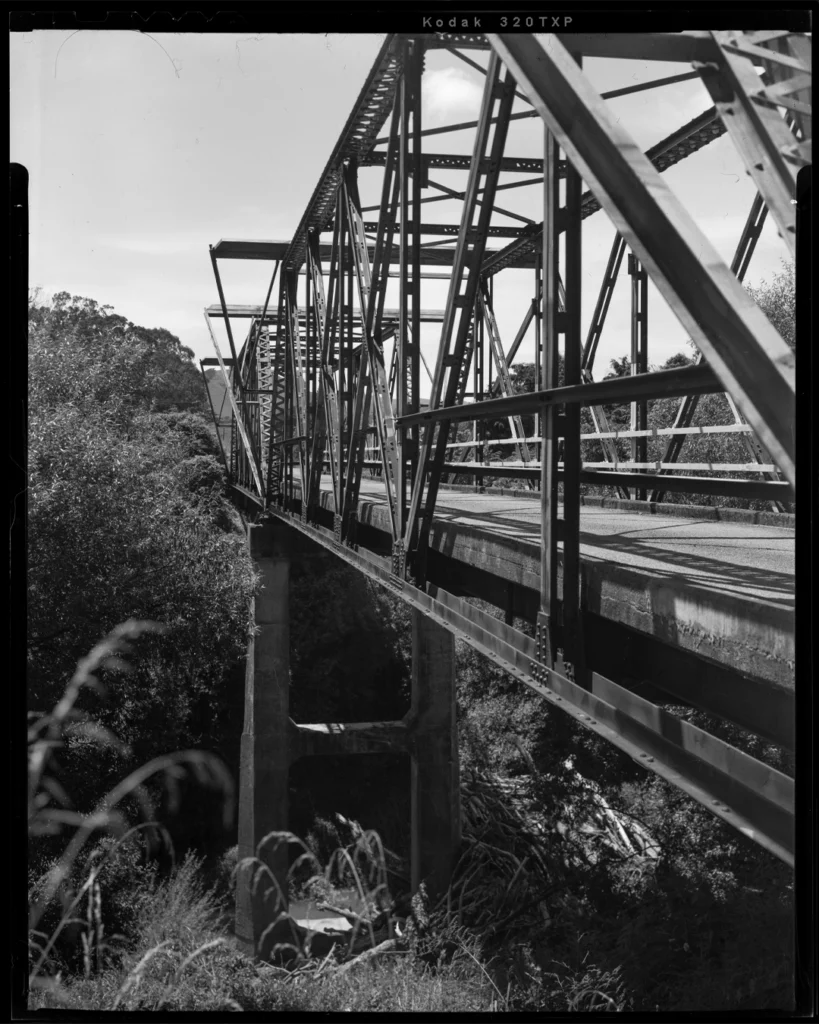 A large format black and white sheet film photograph of a bridge over a river. The river bank is steep and covered in foliage. The bridge is steel and has many struts.