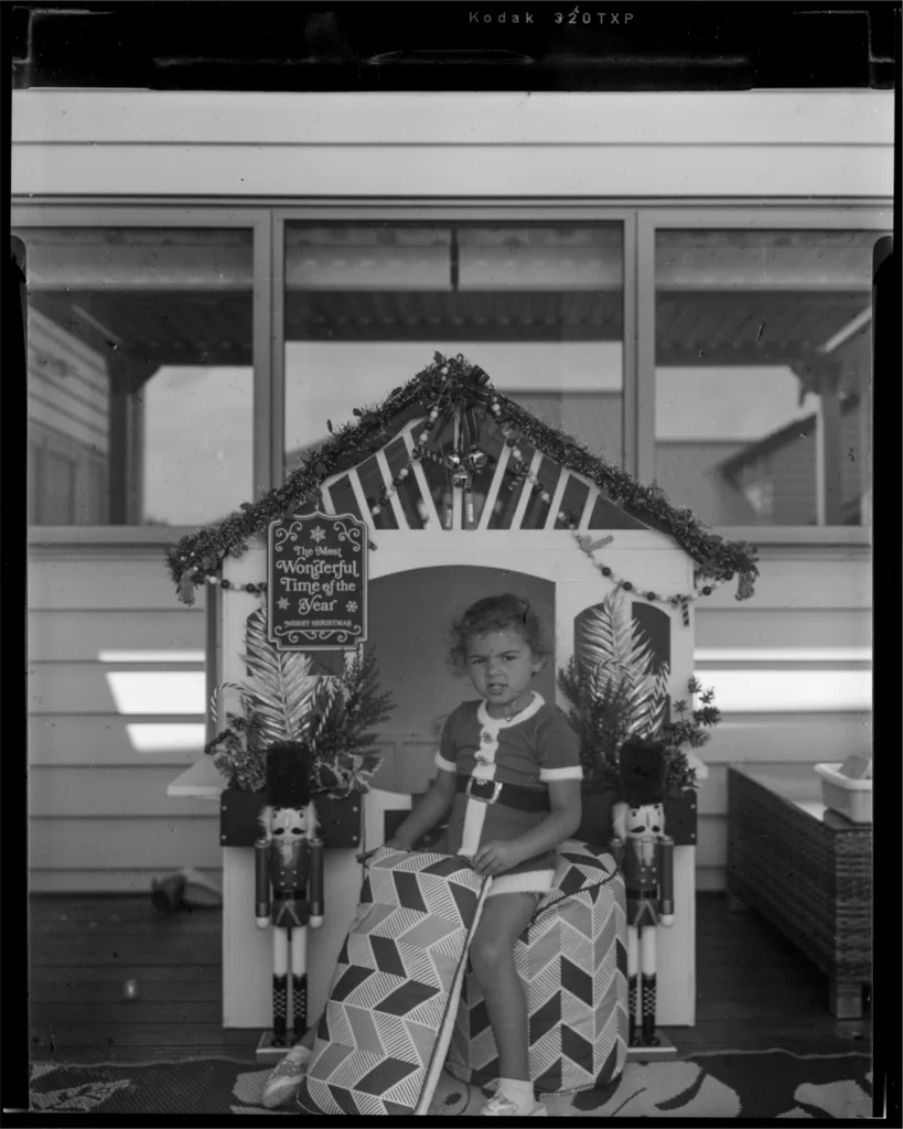A large format black and white sheet film photograph of a young girl sitting posed in front of her playhouse. The playhouse is decorated with holly, and ornamental nutcrackers stand guarding the front entrance.