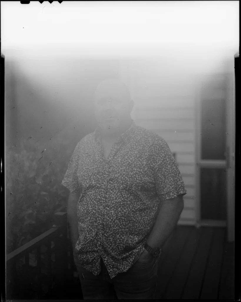 A large format black and white sheet film photograph of a man stood on the deck of a house. He wears a button up short sleeve shirt. A cast of white is beaming in from the top of the photograph.